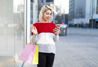 聪明的和年轻的消费者女人站时尚商店窗口显示持有纸购物袋和使用智能手机在阳光明媚的一天在户外年轻的消费者女人站时尚商店窗口显示持有纸购物袋和使用智能手机