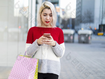 聪明的和年轻的消费者女人站时尚商店窗口显示持有纸购物袋和使用智能手机在阳光明媚的一天在户外年轻的消费者女人站时尚商店窗口显示持有纸购物袋和使用智能手机