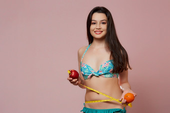 年轻的美丽的纤细的亚洲女孩泳衣与橙色一个手和苹果的其他和测量磁带他的手粉红色的孤立的背景