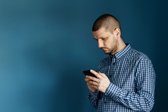 高加索人男人。穿衬衫站前面的蓝色的背景墙使用聪明的电话移动发送消息短信发短信浏览互联网前面视图