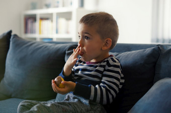 肖像一个小三个年老高加索人男孩小孩子孩子坐着的沙发床上首页持有巧克力糖果手吃而看舔手指口一边视图