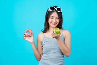 有吸引力的亚洲年轻的女人持有和选择之间的磁盘甜甜圈绿色苹果水果孤立的蓝色的颜色背景重量损失和避免垃圾食物为节食和健康的