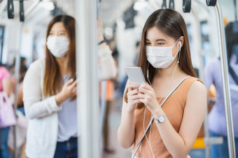 年轻的亚洲女人乘客穿外科手术面具和听音乐通过聪明的移动电话地铁火车当旅行大城市科维德爆发感染和流感大流行概念