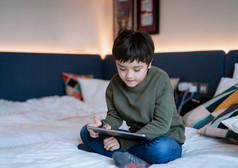 孩子看卡通和聊天与朋友平板电脑快乐男孩坐着床上玩游戏在线数字垫肖像孩子放松首页他的床上房间周末