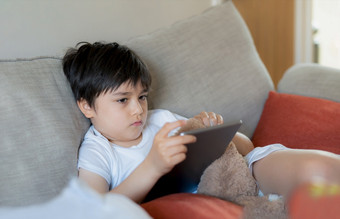 年轻的男孩持有平板电脑看出深认为不开心孩子与伤心脸坐着独自一人沙发孤独的孩子玩与软玩具和玩游戏数字垫首页