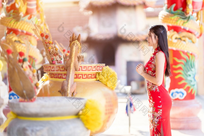 美丽的亚洲年轻的女人穿红色的传统的中国人旗袍装饰站为祈祷佛雕像为中国人新一年节日中国人神社泰国
