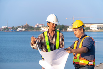 两个工程师站在穿安全头盔站的码头和持有的蓝图和咨询的计划的建设概念非常高效。建设管理