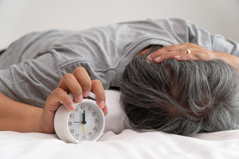 亚洲中年男人。持有白色报警时钟和他的脸显示无聊和感觉坏他的问题睡眠障碍的概念痛苦失眠