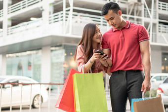 快乐年轻的夫妇<strong>购物</strong>者走的<strong>购物街</strong>对和持有色彩斑斓的<strong>购物</strong>袋手和使用智能手机为检查促销活动概念出售和黑色的星期五<strong>购物</strong>