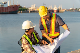 两个工程师站在穿安全头盔站的码头和持有的蓝图和咨询的计划的建设概念非常高效。建设管理