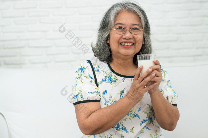 亚洲高级女人持有玻璃牛奶而放松沙发生活房间为退休健康上了年纪的女人喝玻璃牛奶维护她的幸福概念健康和健康的