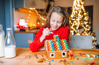 可爱的小女孩装修姜饼房子与釉美丽的厨房与灯和圣诞节树表格与蜡烛快乐圣诞节和快乐假期小女孩使圣诞节姜饼房子的厨房