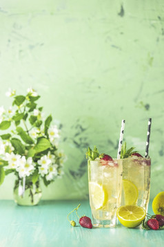 两个眼镜冷冰冷的让人耳目一新喝与柠檬和草莓服务与酒吧工具绿色木表格与白色盛开的花新鲜的鸡尾酒饮料与冰水果和草装饰