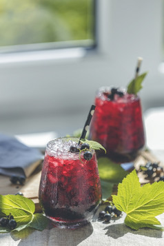 两个玻璃冷冰黑色的醋栗汁与成熟的浆果和绿色叶子表格阳光明媚的房间附近窗口与花园外
