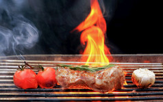 烤牛肉牛排迷迭香西红柿和大蒜燃烧的木炭烧烤与烟和火火焰黑色的背景
