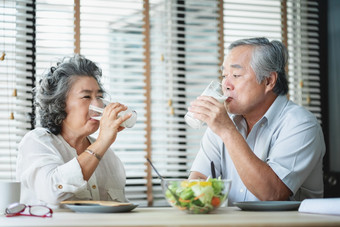 微笑亚洲高级男人。和女人坐着表格喝眼镜牛奶和看每一个其他快乐的上了年纪的夫妇享受健康的沙拉食物而有早餐首页