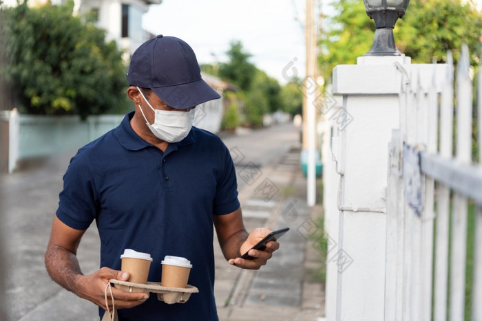 非洲美国交付男人。蓝色的统一的和保护脸面具使用移动电话和持有杯热喝而等待客户端前面房子在冠状病毒检疫
