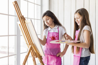 关闭两个女孩站附近窗口绘画画架与<strong>油漆刷</strong>