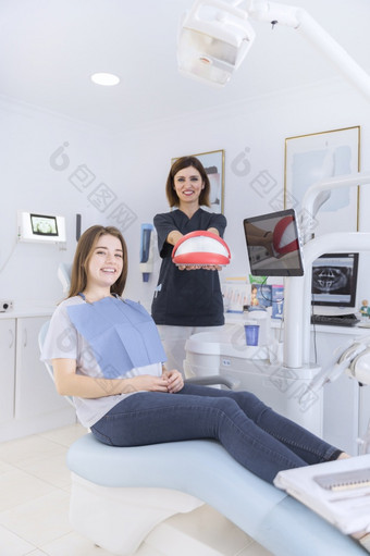 快乐女坐着牙科椅子前面牙医持有牙齿模型