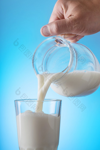 手倒牛奶从壶成玻璃蓝色的背景的概念健康的自然食物手倒牛奶从壶成玻璃蓝色的背景