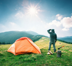 男人。山附近橙色帐篷看起来成的距离男人。罩站附近橙色帐篷和看起来的明亮的太阳附近的是柴火和夏天景观的概念隐私和旅行男人。山附近橙色帐篷看起来成的距离