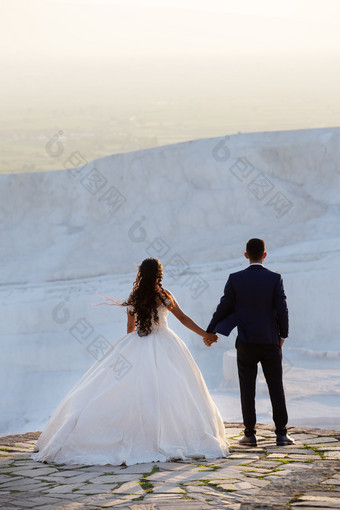 年轻的婚礼夫妇站在悬崖持有手和看起来成的距离男人。和女人婚礼服装情人站的背景白色棉花堡石灰华的视图从的回来概念爱和的关系年轻的婚礼夫妇站在悬崖持有手和看起来成的距离