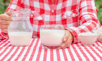 女人网纹红色的衬衫持有出玻璃牛奶的表格红色的桌布的桌布壶牛奶和酸奶油采购为广告项目女人红色的衬衫持有出玻璃牛奶