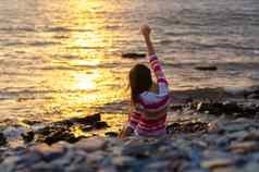 女孩坐在海滨抛出石头水提高了手晚上日落