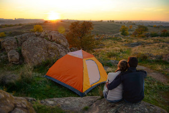 年轻的夫妇坐着帐篷看美女日落山冒险家庭旅行
