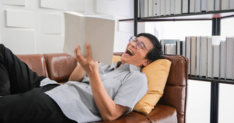 高级亚洲男人。阅读书笑沙发生活房间首页肖像亚洲上了年纪的男人。放松幸福读杂志首页