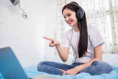 亚洲快乐年轻的女人穿耳机听音乐首歌看网站购物在线移动PC床上首页