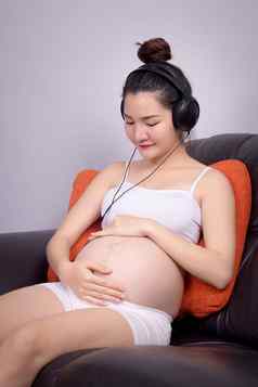 肖像怀孕了亚洲女人坐着沙发首页听音乐耳机美丽的亚洲女模型