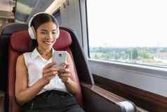 现代人城市生活方式年轻的城市女人电话应用程序无线耳机听音乐玩视频游戏在线亚洲女孩享受火车旅行业务类座位