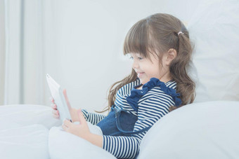 可爱的女孩享受看卡通聪明的平板电脑可爱的娃娃坐着床上卧室首页