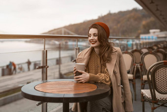快乐年轻的女孩坐着在户外街餐厅阳台微笑持有咖啡杯子肖像时尚的年轻的女人穿秋天外套红色的贝雷帽在户外