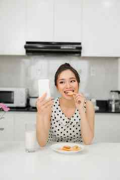 年轻的微笑女人吃饼干微笑智能手机健康的早餐