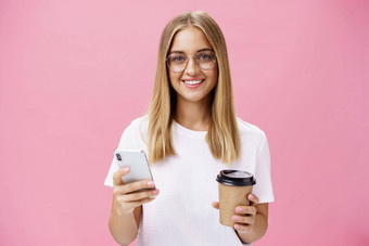 愉快的友善的女孩微笑相机持有纸杯咖啡智能手机肖像快乐的不错的女人喝早....喝摆姿势的意见咖啡馆互联网粉红色的墙