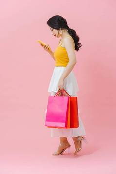 快乐年轻的女人持有购物袋移动电话粉红色的背景