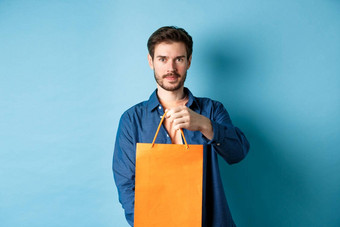 英俊的有胡子的男人。拉伸手橙色<strong>购物</strong>袋使礼物站蓝色的背景