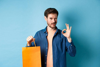 现代男人。休闲装显示购物袋标志眨眼相机推荐商店站蓝色的背景
