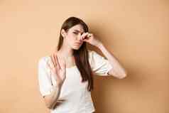 不高兴女人插头鼻子坏气味显示停止手势皱着眉头厌恶关闭鼻孔厌恶站米色背景