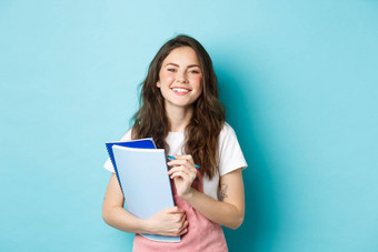 快乐年轻的女学生持有笔记本电脑课程微笑相机站春天衣服蓝色的背景