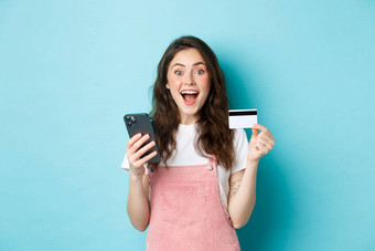 惊讶快乐年轻的女人显示塑料信贷卡智能手机惊讶相机购物在线春天折扣蓝色的背景