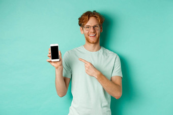 有吸引力的年轻的男人。红色的胡子头发指出手指空白智能手机屏幕显示在线促销活动应用程序微笑相机绿松石背景