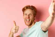 兴奋红色头发的人男人。旅游采取自拍显示竖起大拇指持有相机拉伸手站粉红色的背景
