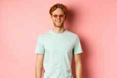 旅游假期概念快乐的红色头发的人有胡子的男人。太阳镜t恤微笑快乐相机站粉红色的背景