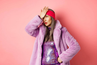 时尚购物概念生气高级女人卷眼睛使facepalm指蹩脚的站时尚的紫色的冬天外套闪闪发光的衣服粉红色的背景