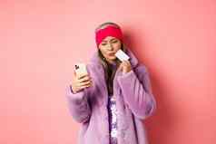 在线购物时尚概念时尚的亚洲夫人紫色的人造皮毛外套优柔寡断智能手机屏幕持有塑料信贷卡粉红色的背景