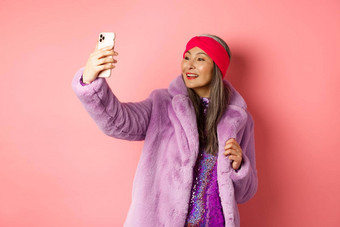时尚概念美丽的亚洲高级夫人采取自拍智能手机摆姿势聚会，派对闪闪发光的衣服紫色的人造外套站粉红色的背景