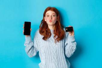 兴奋红色头发的人女孩显示移动电话屏幕信贷卡展示在线商店应用程序站蓝色的背景